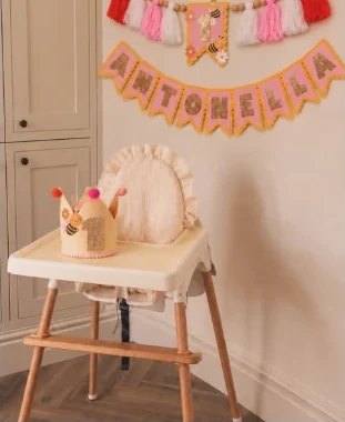 Guirnalda rosada con el nombre Antonella y una coronita con abejas sobre una sillita de comer